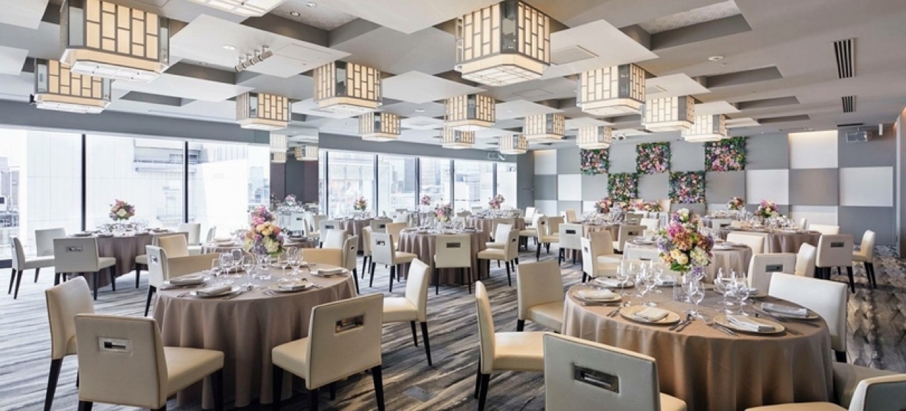THE GRAND GINZAのバンケット「THE GRAND GALLERY」。ラウンドテーブルに白い椅子と花装飾、格子柄の照明と銀座を望む大きな窓が並ぶ宴会場内観